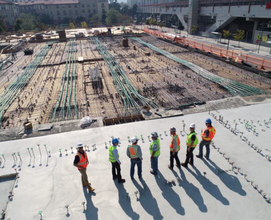 Seven people standing on site overlooking the construction of a building. They are all wearing high viz jackets and hard hats in different colours.