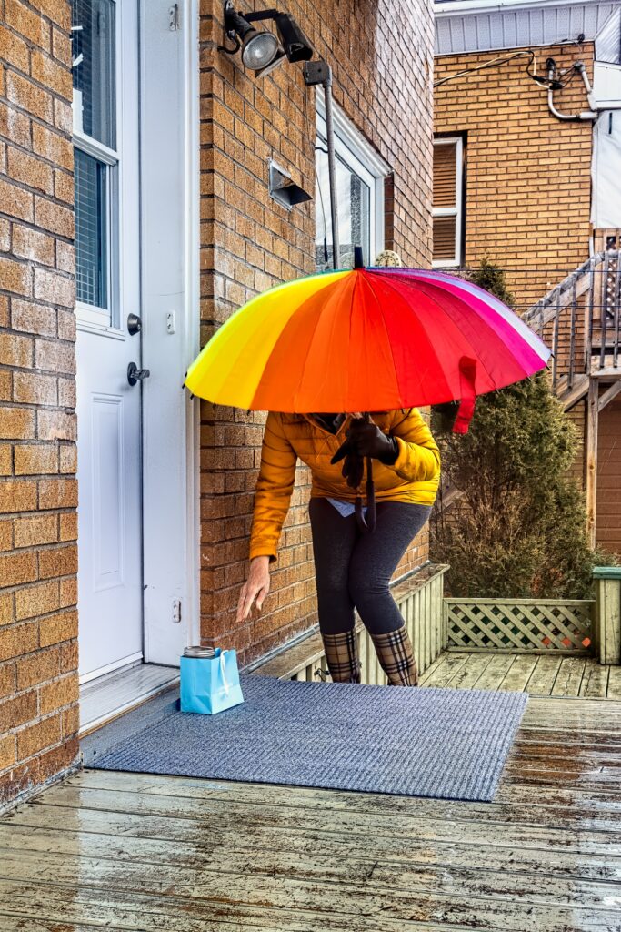 A woman holding a rainbow coloured umbrella in front of a front door, bending down to pick of a gift bag.