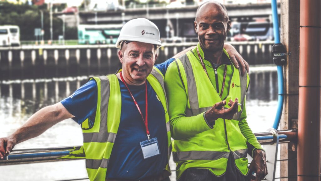 Two men with there arms around each other looking at the camera. They are both wearing high viz vest and the one on the left is wearing a white hard hat. They are leaning on railings in front of a river.
