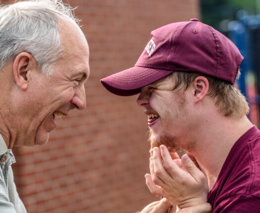 A young man and an older man looking at each other and laughing, they are outside in front of a brick wall and the young man is wearing a cap.