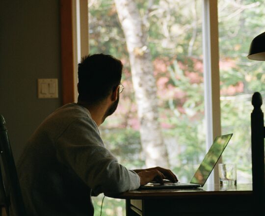 A man set a table on a laptop looking out the window at some trees.